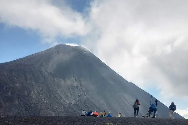 Small groups of hikers resting on dark sand at the foot of the steep grey cone