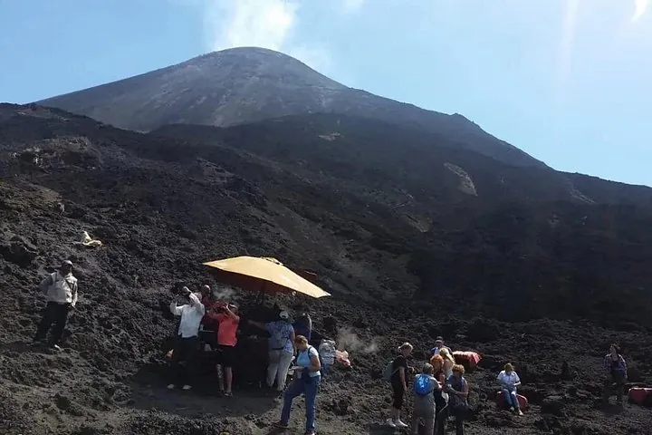 A tour group gathered under an orange umbrella on a lava field below the cone